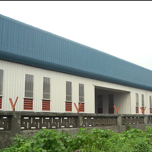 Long industrial building with a blue roof and white walls, surrounded by greenery.