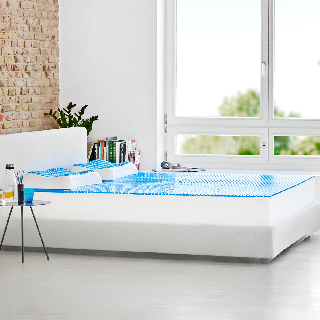 Mattress topper with blue gel layer on a bed in a bright room with a window.
